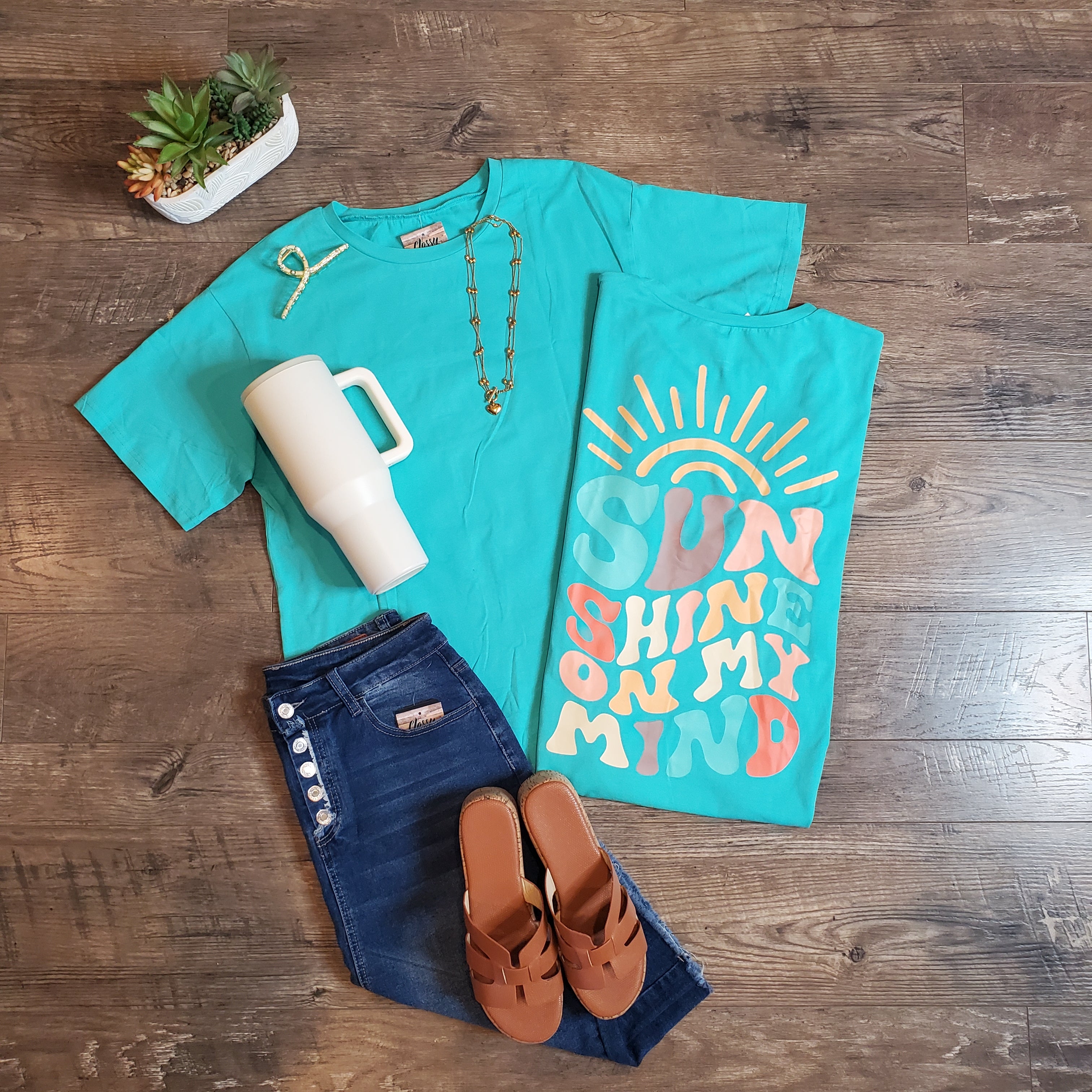 Turquoise t-shirt with 'Shine on my mind' design, blue jean skirt, brown sandals, white mug, and necklace on a wooden surface.