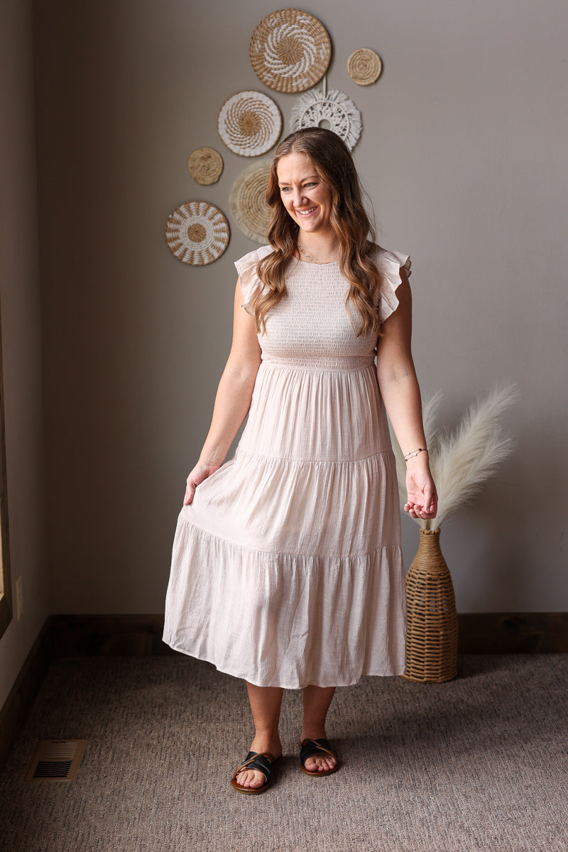 Woman in a beige dress standing in a room with decorative wall art and a vase.