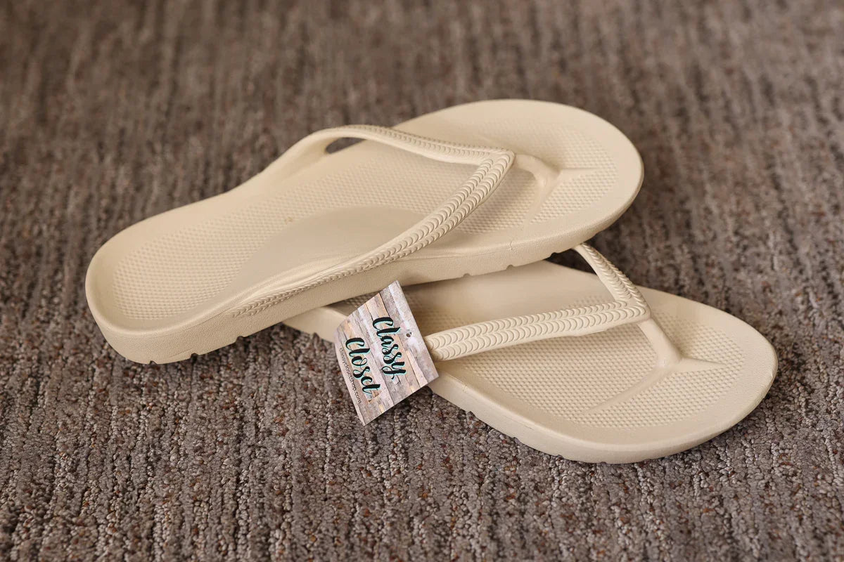 Pair of beige flip-flops with a tag on a brown textured surface