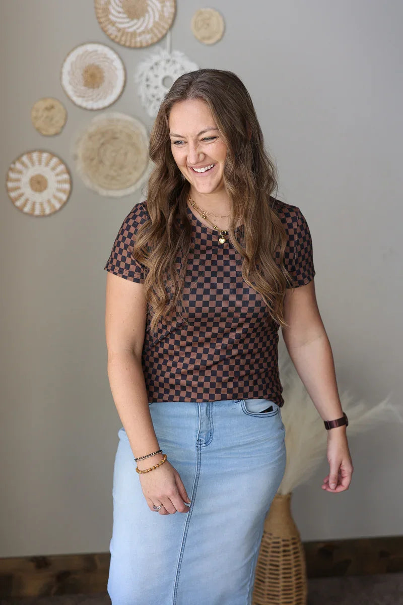 Woman wearing a checkered top and blue jean skirt standing in front of decorative wall hangings.