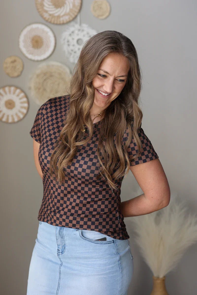 Woman wearing a checkered top and light blue jean skirt against a neutral wall with decorative elements.