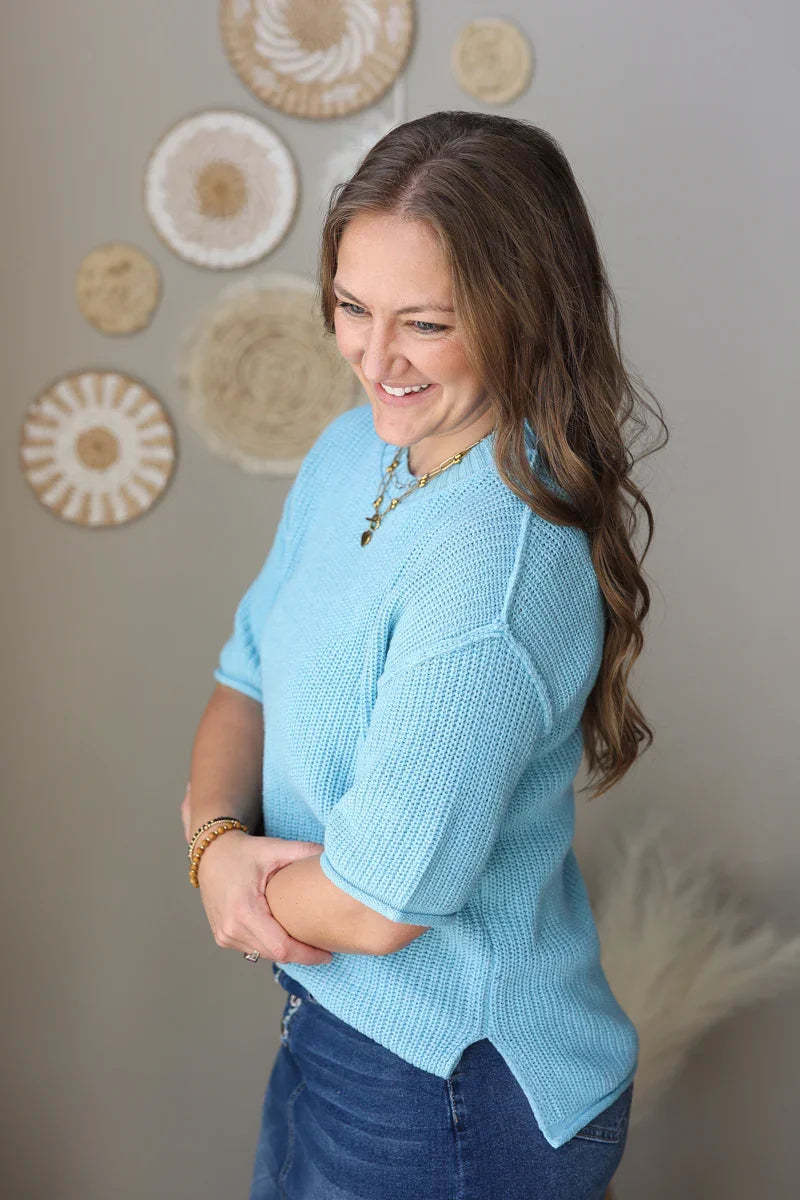 Woman wearing a light blue sweater and jean skirt standing against a neutral wall with decorative elements.