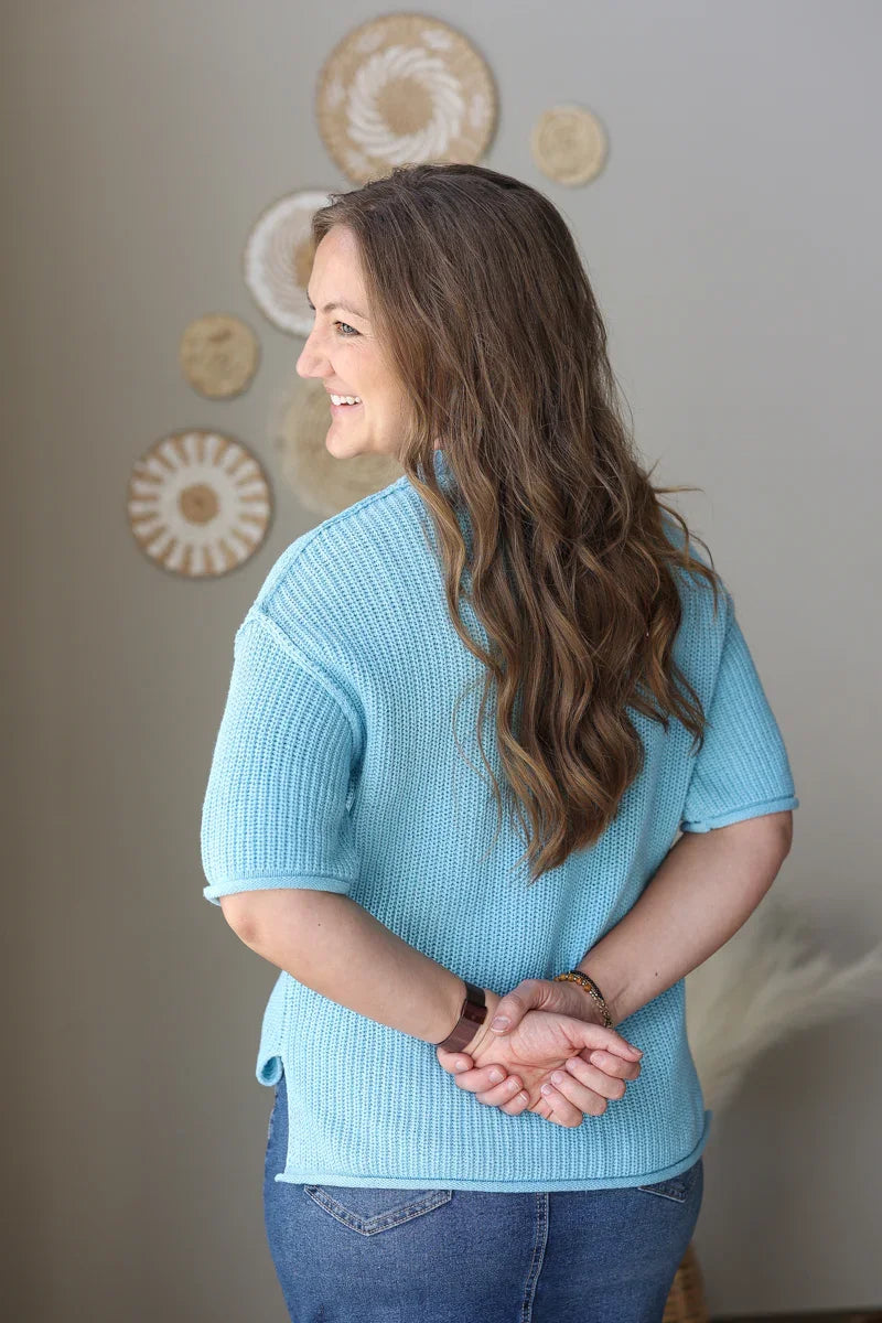 Woman in a blue sweater and jeans holding a mug and a bag, standing against a wall with decorative items.