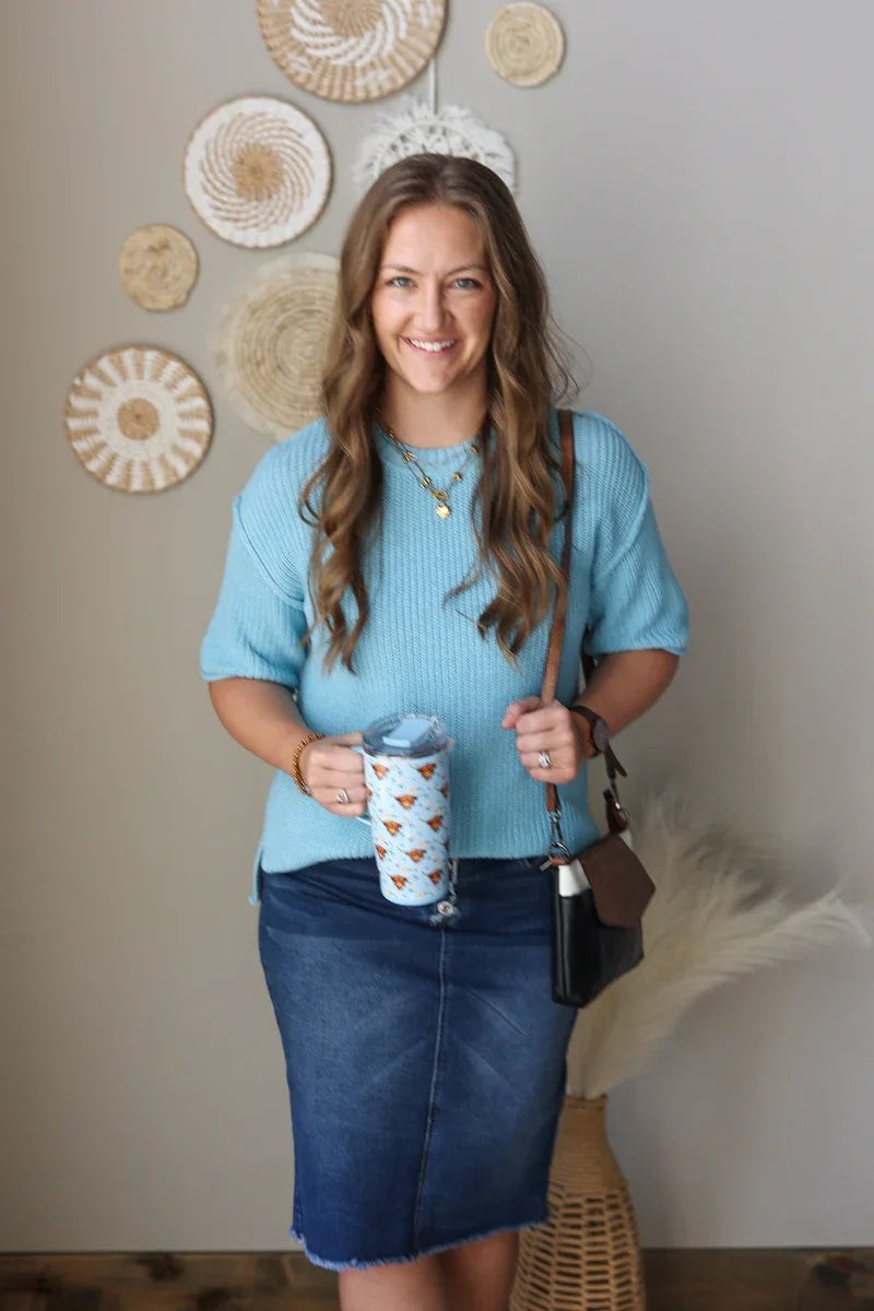 Woman in a blue sweater and denim skirt holding a mug with decorative elements on the wall.