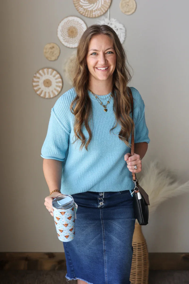 Woman in a blue sweater and denim skirt holding a coffee cup and purse indoors.