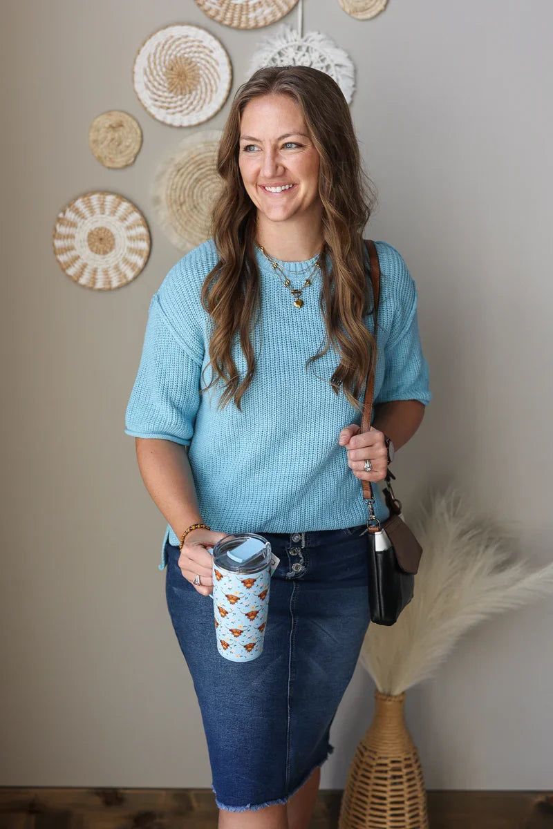 Woman in a blue sweater and denim skirt holding a coffee cup and a phone case, with decorative wall art in the background.
