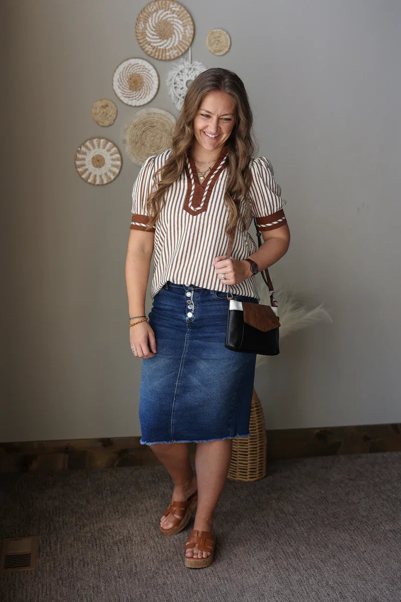 Woman wearing a striped shirt and denim skirt standing indoors with decorative wall art in the background.