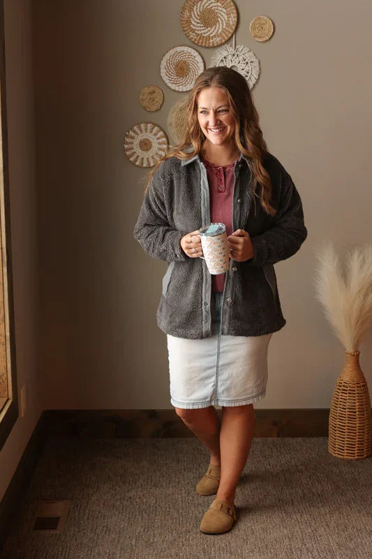Woman wearing a charcoal sherpa shacket, light wash denim skirt, and thermal henley top, holding a mug in a room with decorative wall art