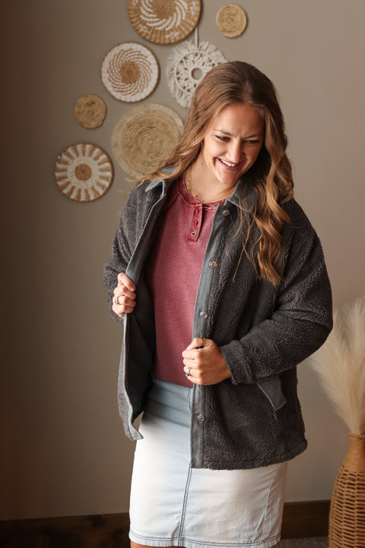Woman wearing a dark jacket over a thermal henley top and light wash denim skirt, standing in a room with decorative wall art.