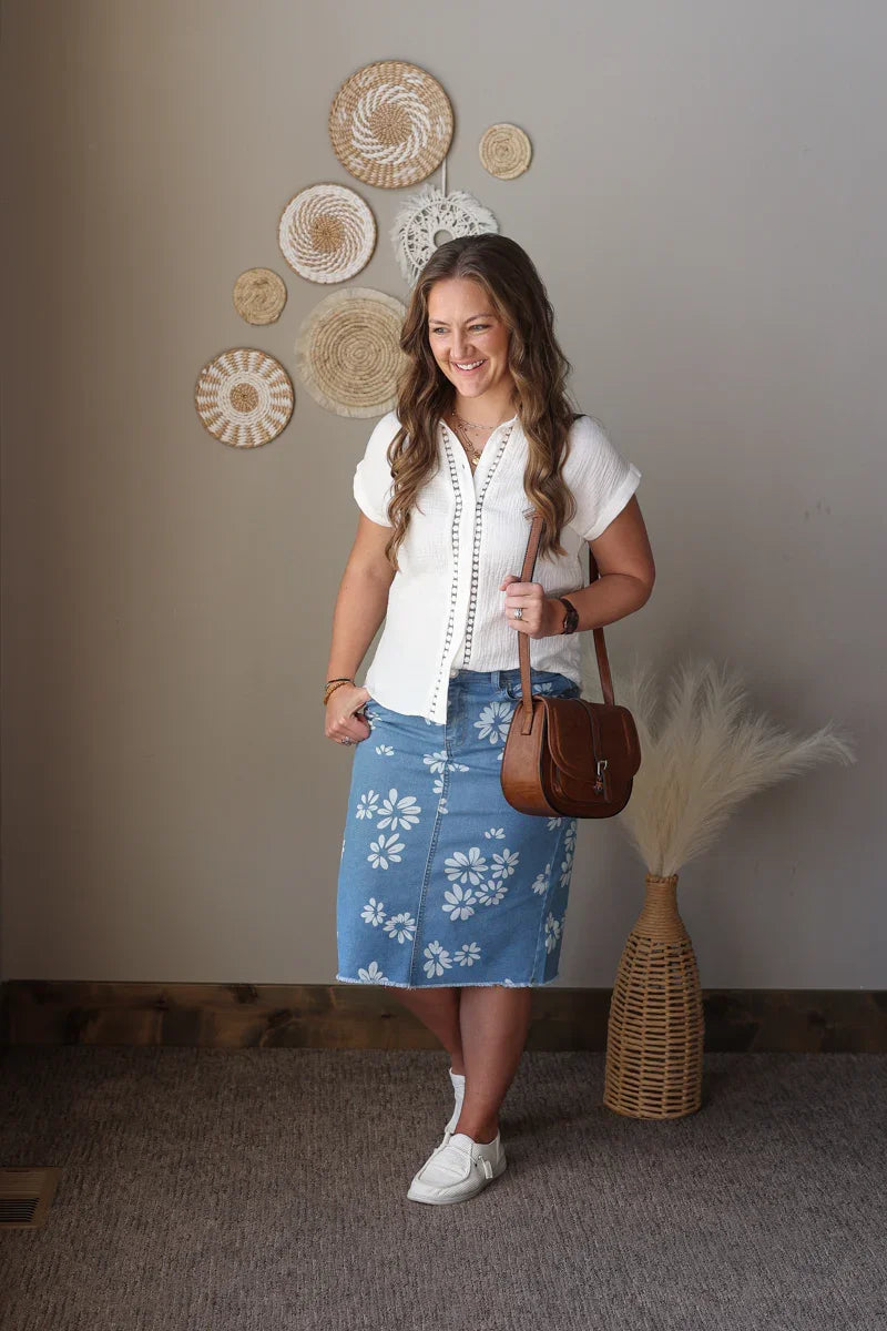 Woman in a white blouse and blue floral skirt holding a brown handbag, standing in a room with decorative wall art.