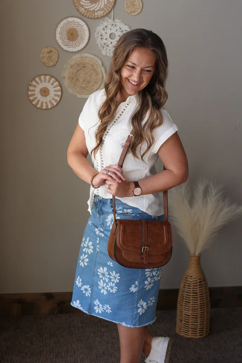 Woman in a white top and blue floral skirt holding a brown handbag, standing against a wall with decorative plates.