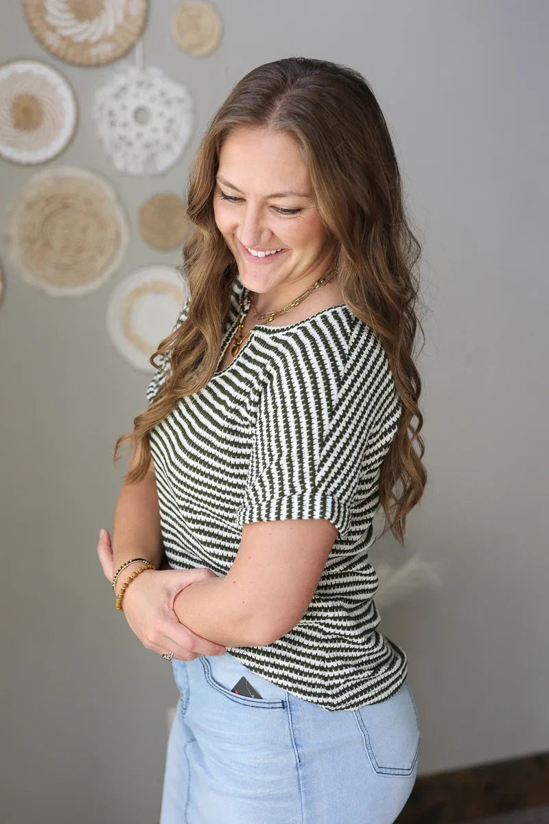Woman wearing a dark green and white striped shirt and blue jean skirt standing against a neutral wall with decorative items.