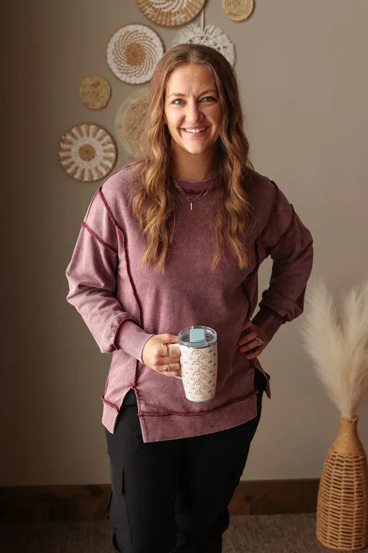 Woman holding a mug in a room with decorative wall art and a vase. Woman wearing Burgundy Mineral Wash Side Slit Sweatshirt with relaxed fit, styled casually with joggers, perfect for comfortable everyday wear