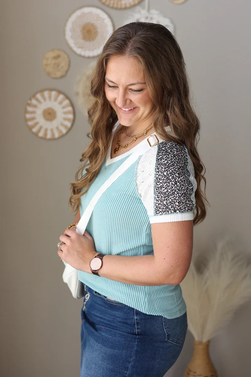 Woman wearing a light blue top with a white crossbody over it, standing against a neutral wall.