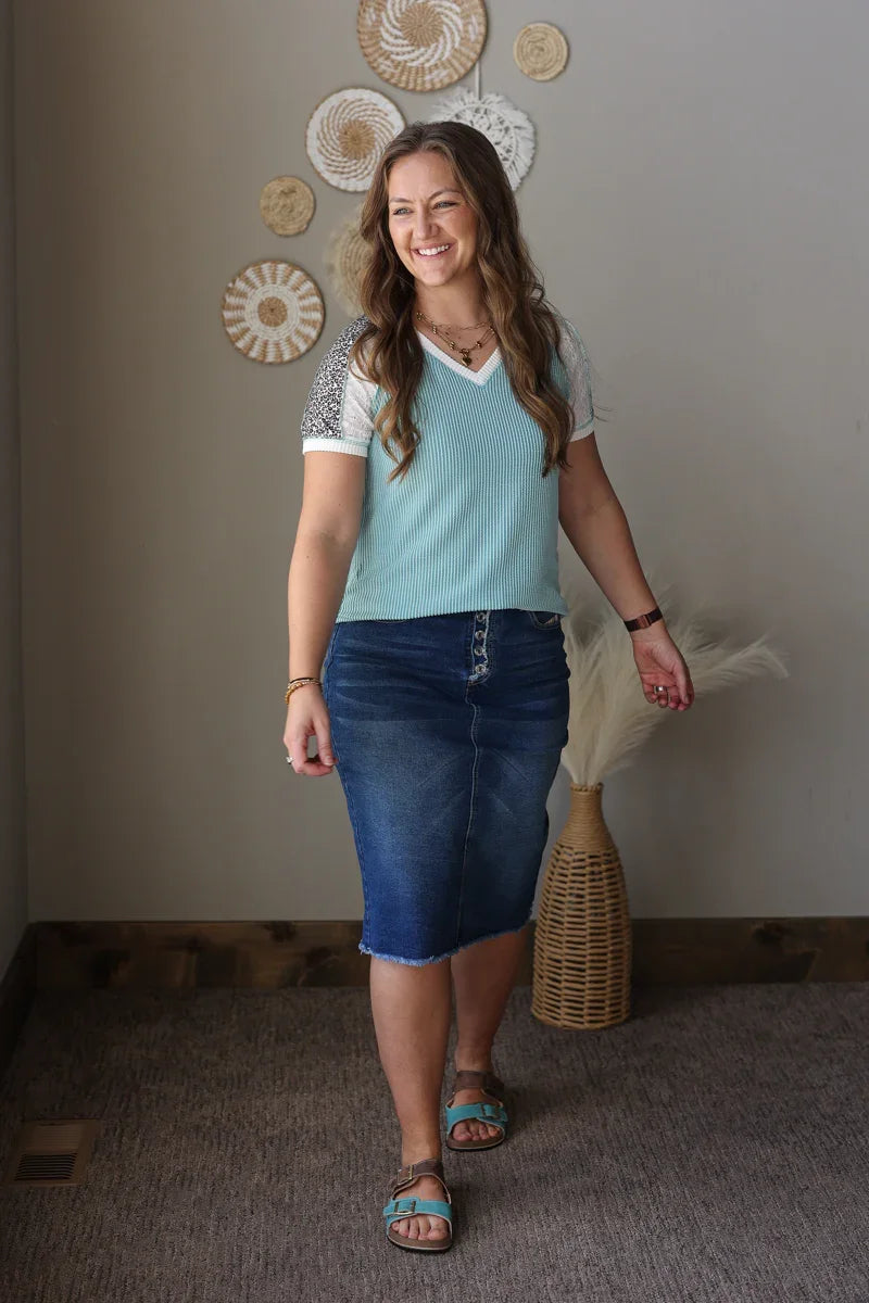 Woman wearing a light blue top and denim skirt standing in a room with decorative wall art.