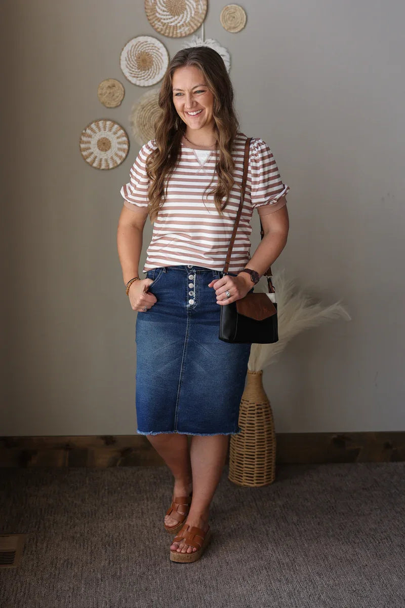 Woman wearing a striped brown shirt and denim skirt standing indoors with decorative wall art in the background.