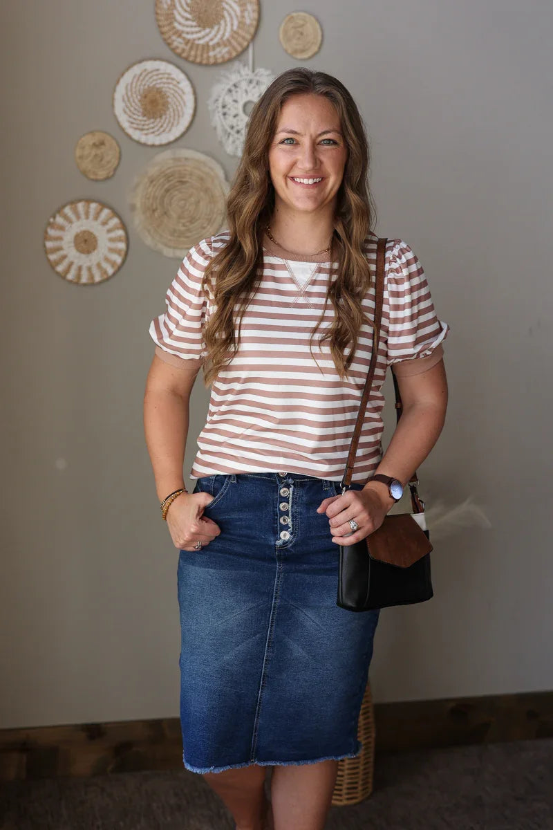 Woman wearing a tan mocha striped shirt and denim skirt with a brown bag, standing against a wall with decorative plates.