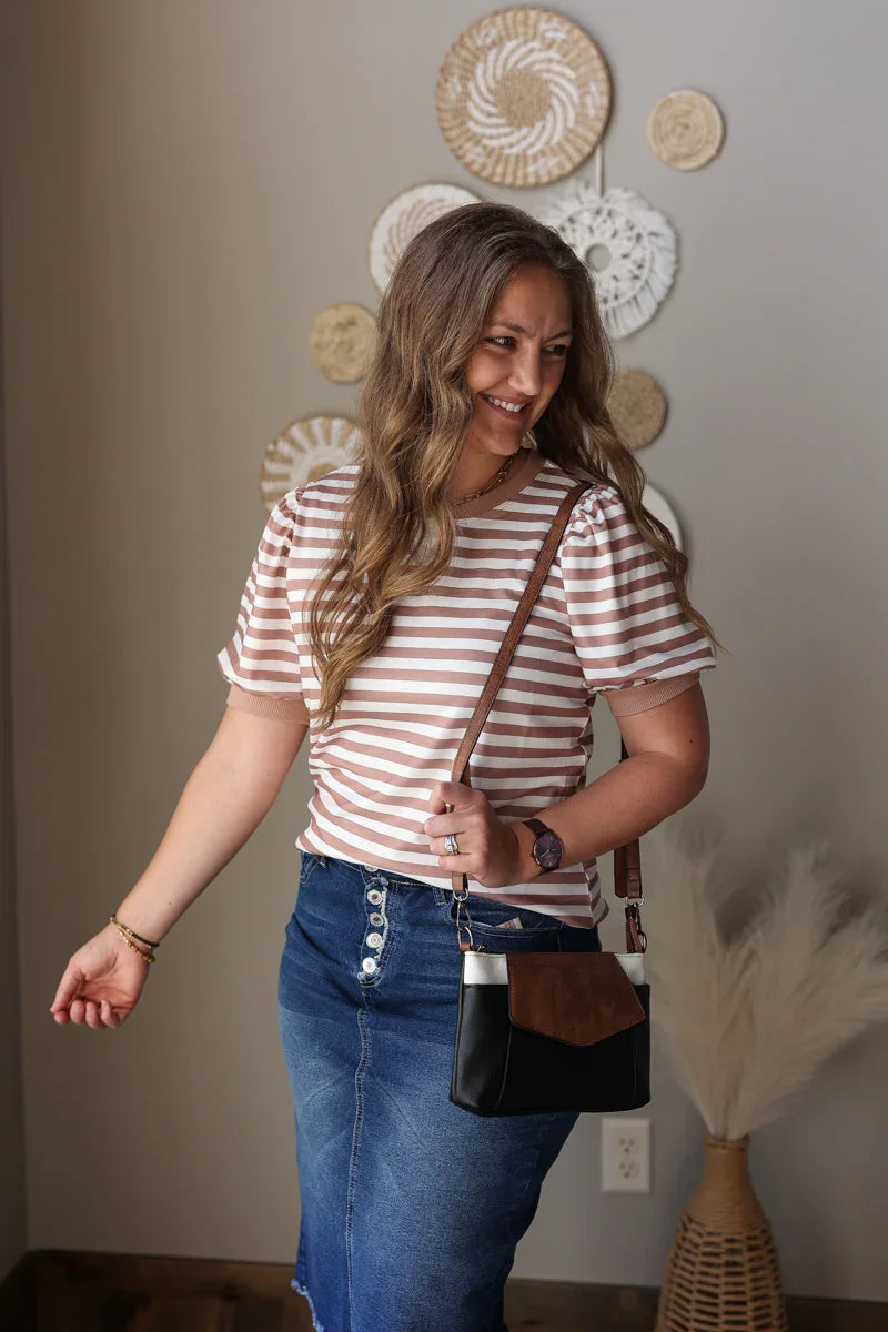 Woman wearing a striped brown shirt and blue jean skirt, holding a brown bag, standing in a room with decorative wall art.