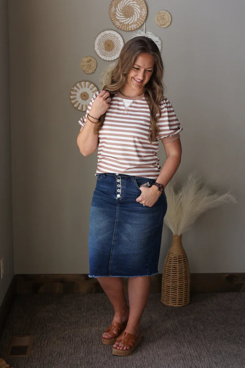 Woman wearing a striped brown shirt and denim skirt standing in a room with decorative elements.