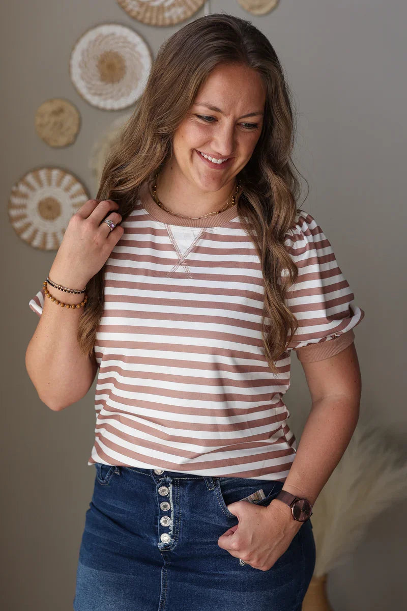 Woman wearing a striped brown shirt and blue jean skirt with decorative wall art in the background