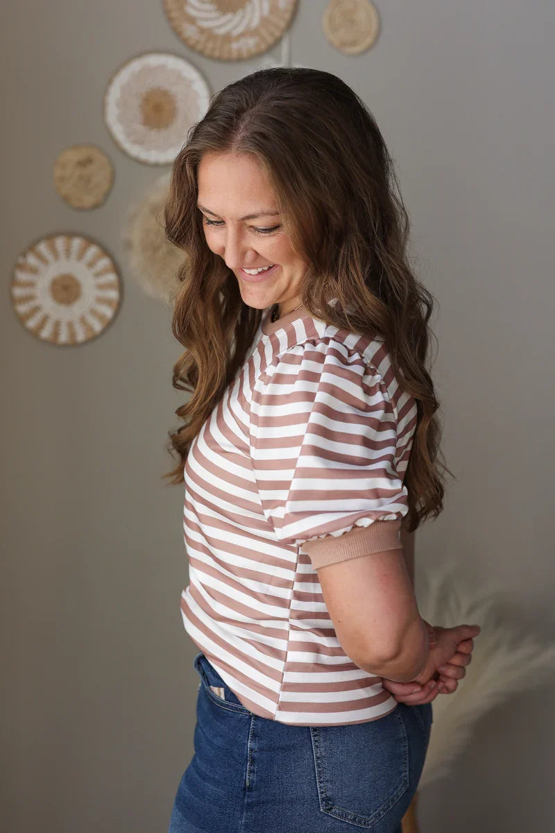 Woman wearing a striped mocha shirt and jean skirt standing against a neutral wall with decorative elements.