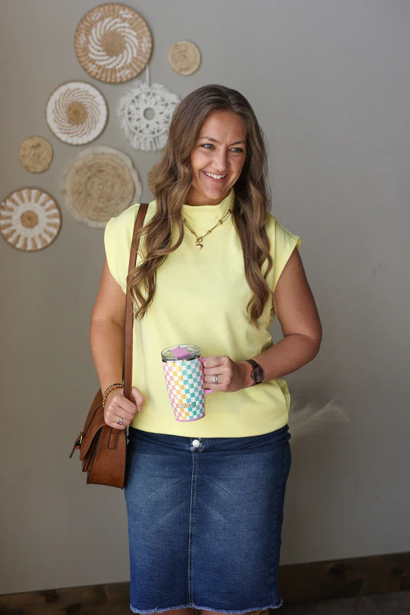Woman in a yellow top and blue skirt holding a colorful cup and brown bag against a wall with decorative plates.