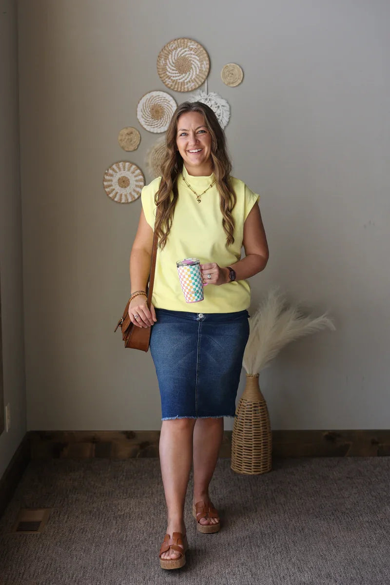 Woman in a yellow top and blue skirt holding a colorful cup, standing in a room with decorative wall art.