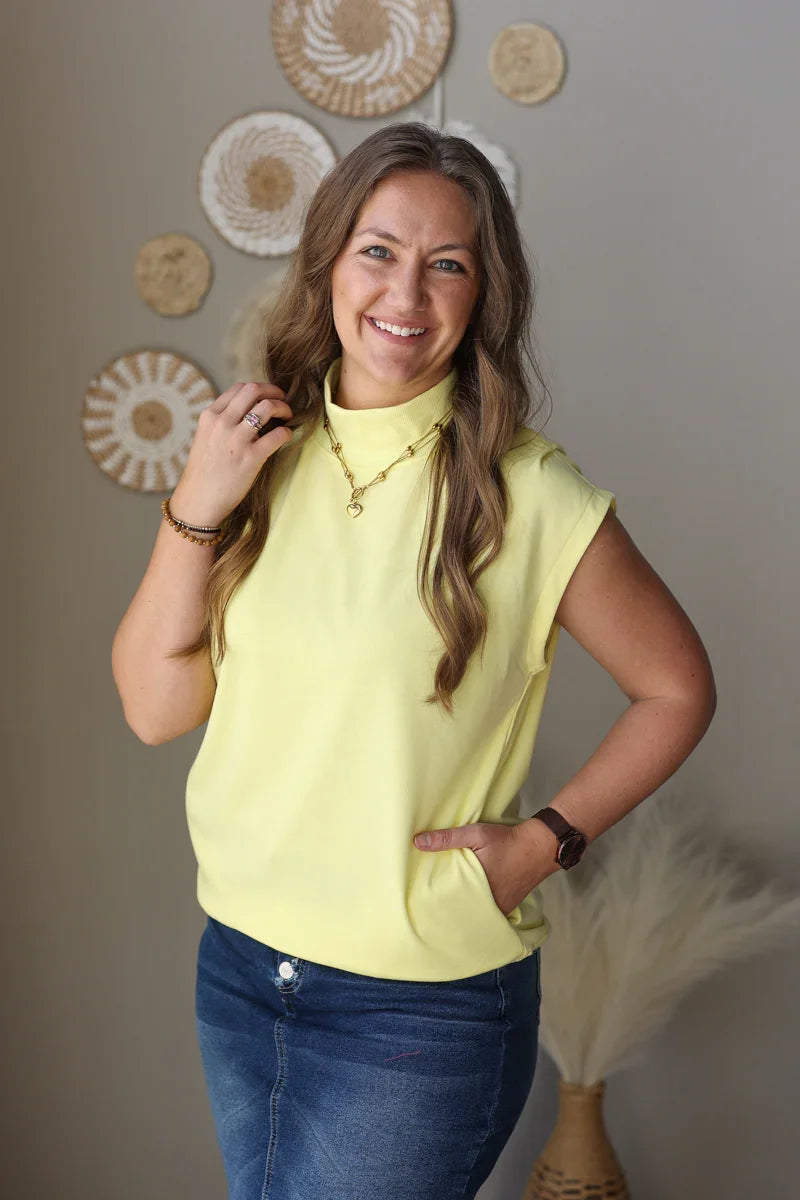 Woman wearing a yellow top and blue jean skirt standing against a neutral wall with decorative elements.