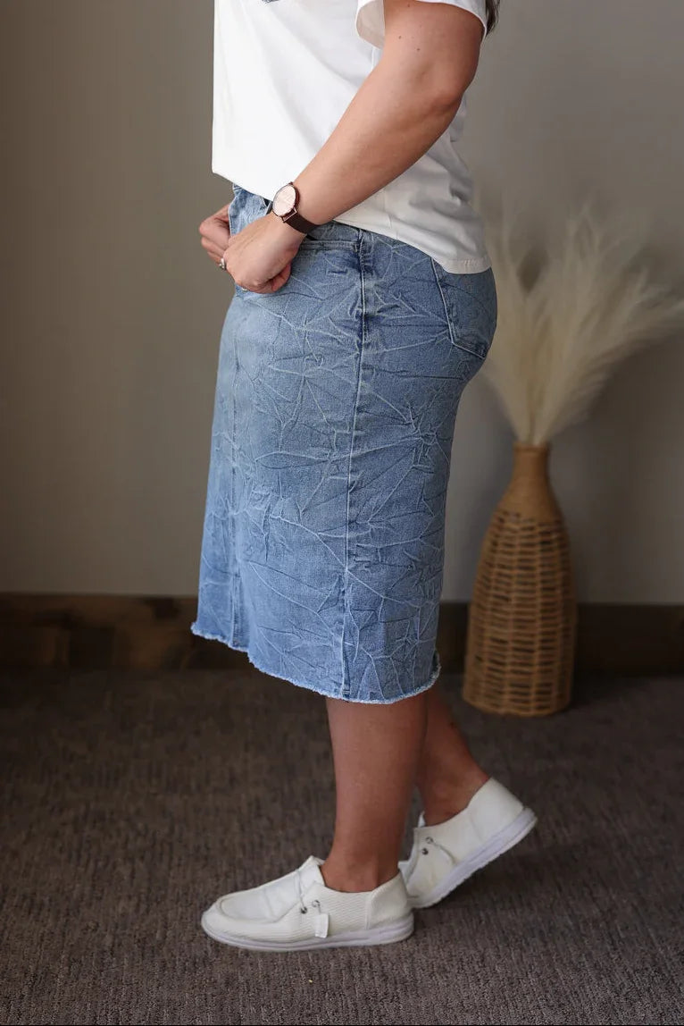 Person wearing a white t-shirt and blue denim skirt with white sneakers, standing indoors.