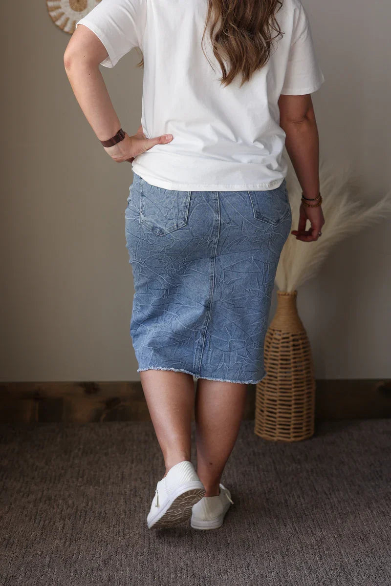 Person wearing a white t-shirt and blue denim skirt standing indoors with a neutral background.