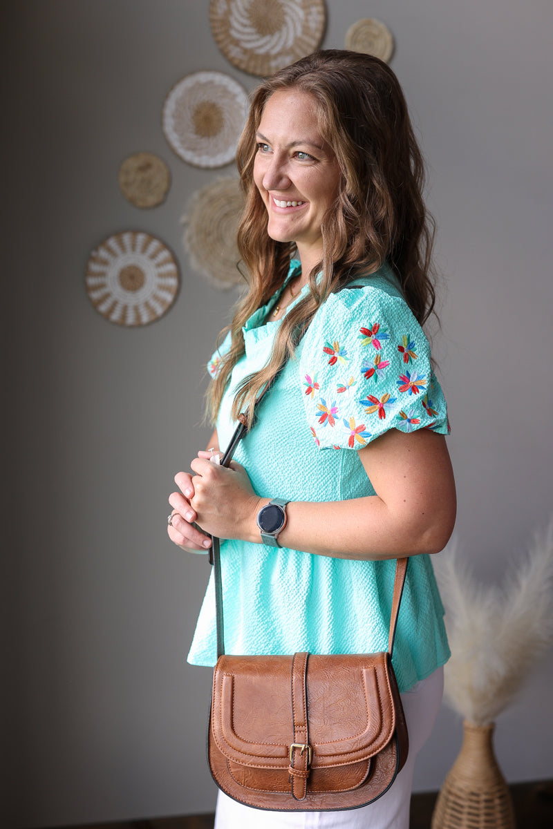 Woman wearing a turquoise floral blouse with a brown leather bag indoors.