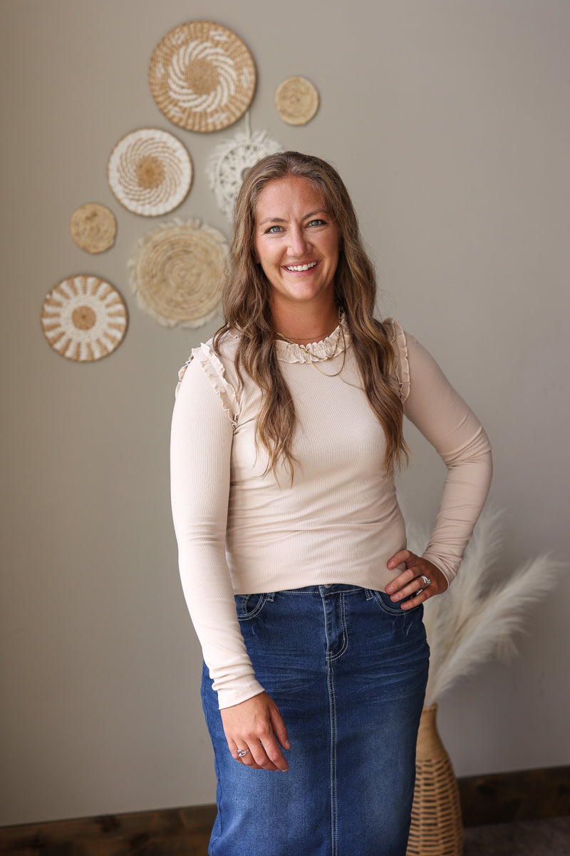 A woman with long light brown hair stands indoors wearing a fitted cream long-sleeve top with lace-like shoulder details and a blue denim skirt. She is posed against a beige wall decorated with woven circular wall pieces, with a pampas grass arrangement in a basket at the lower right.