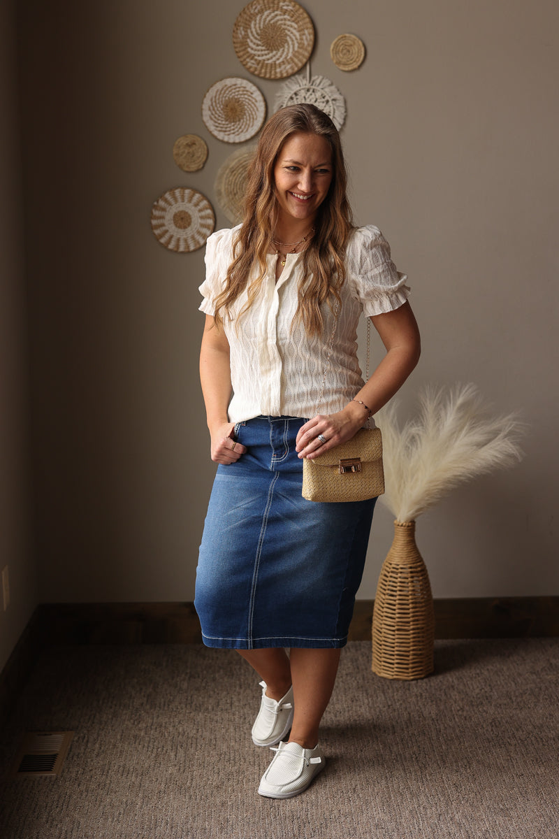 Woman in a white blouse and blue skirt standing in a room with decorative wall art and a vase.