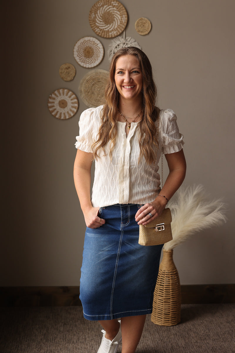 Woman in a white blouse and blue skirt holding a beige bag against a wall with decorative plates.