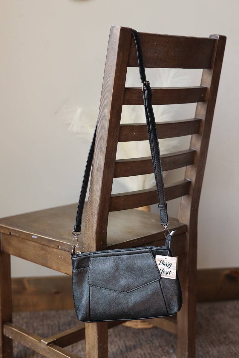 A black faux-leather crossbody handbag with a front pocket and a white 'thing you want' tag, draped over a dark wooden ladder chair.