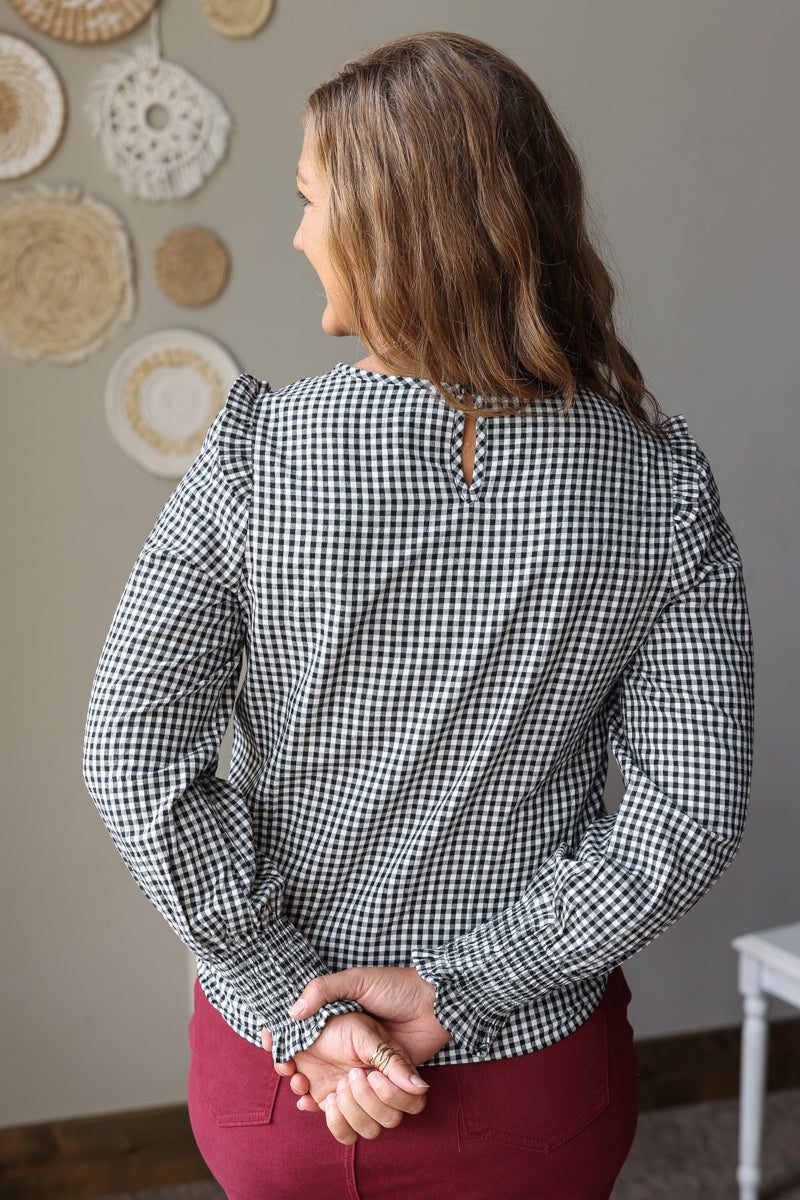 A woman viewed from behind wearing a black and white gingham long-sleeve blouse with ruffled shoulders and a small keyhole back detail, paired with burgundy denim skirt, standing in a decorated indoor setting.