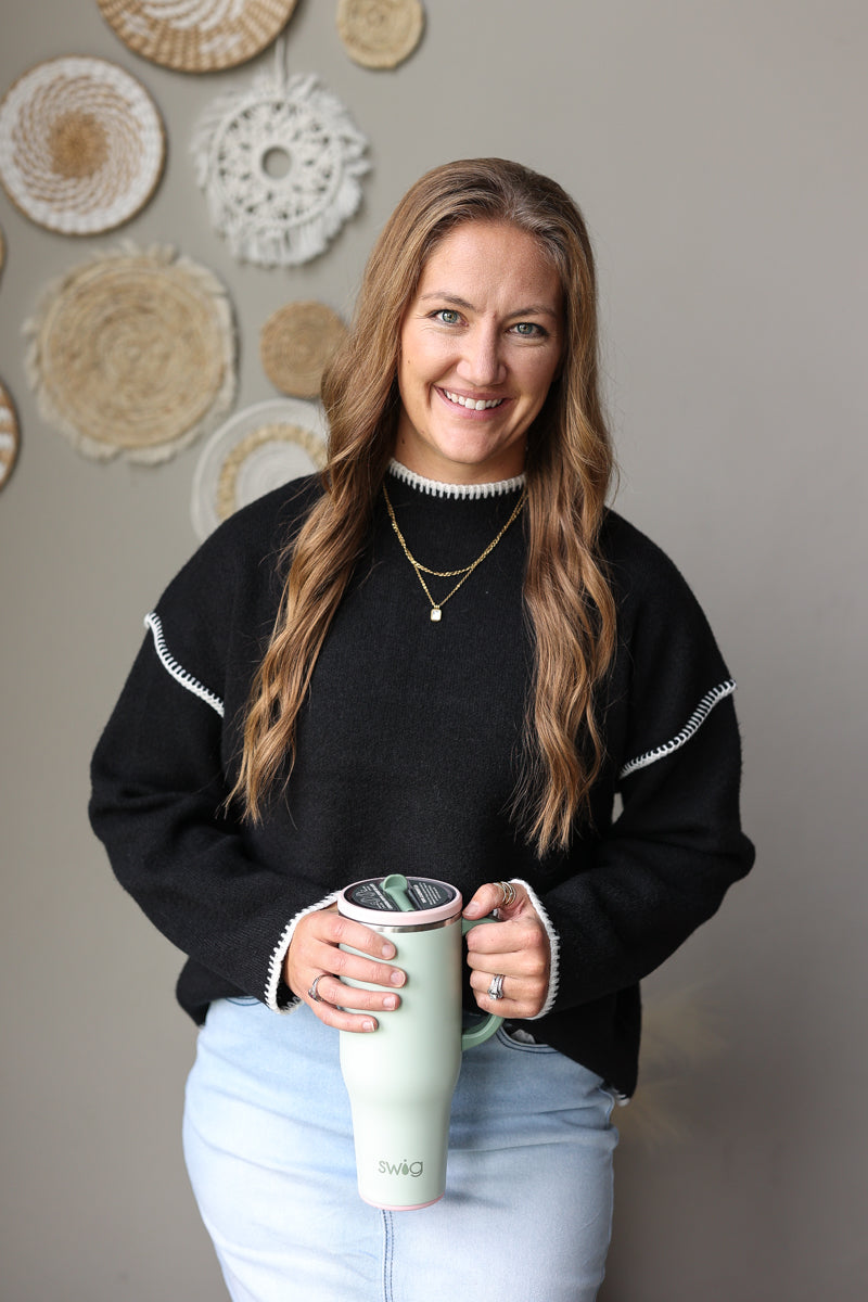 Front view of a black long sleeve knit sweater showing the mock neckline, ribbed cuffs, and contrast stitching.