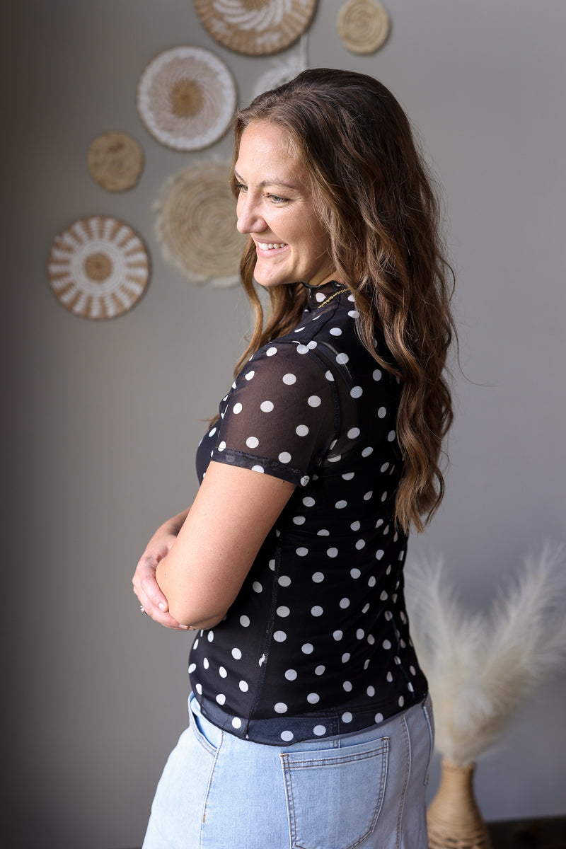 Woman wearing a black polka dot shirt with light blue jean skirt in a room with decorative wall art.