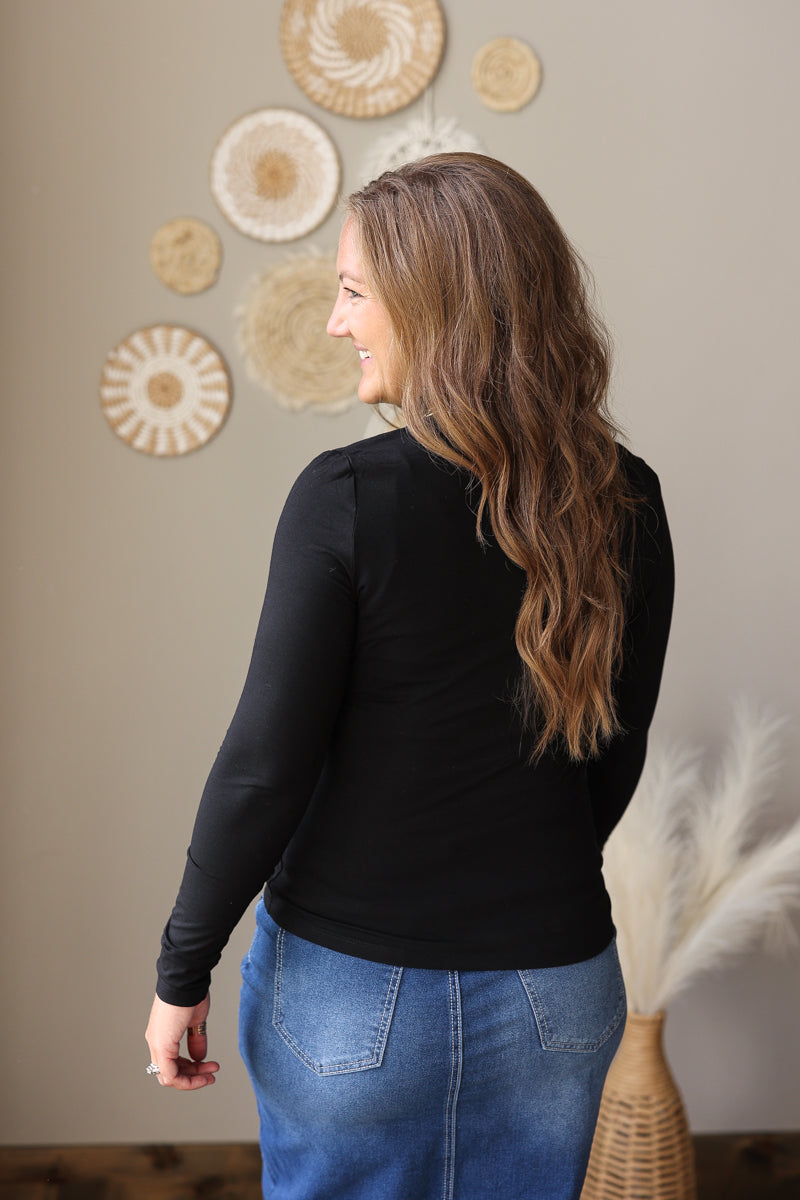 A woman with long wavy brown hair is shown from the back wearing a black long-sleeve top and a blue denim skirt, standing in a room with decorative wall art and a vase of pampas grass.