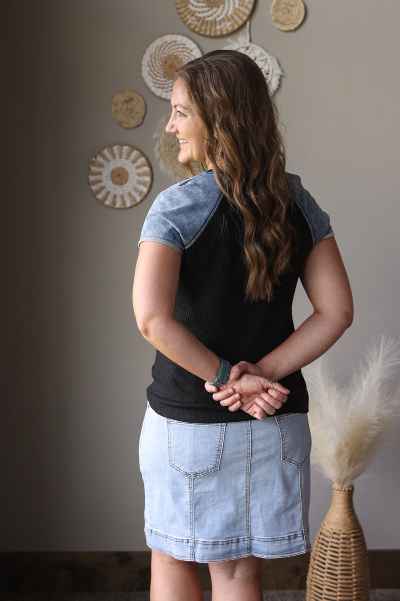 Woman wearing a black top with denim sleeves and skirt in a room with decorative wall art.