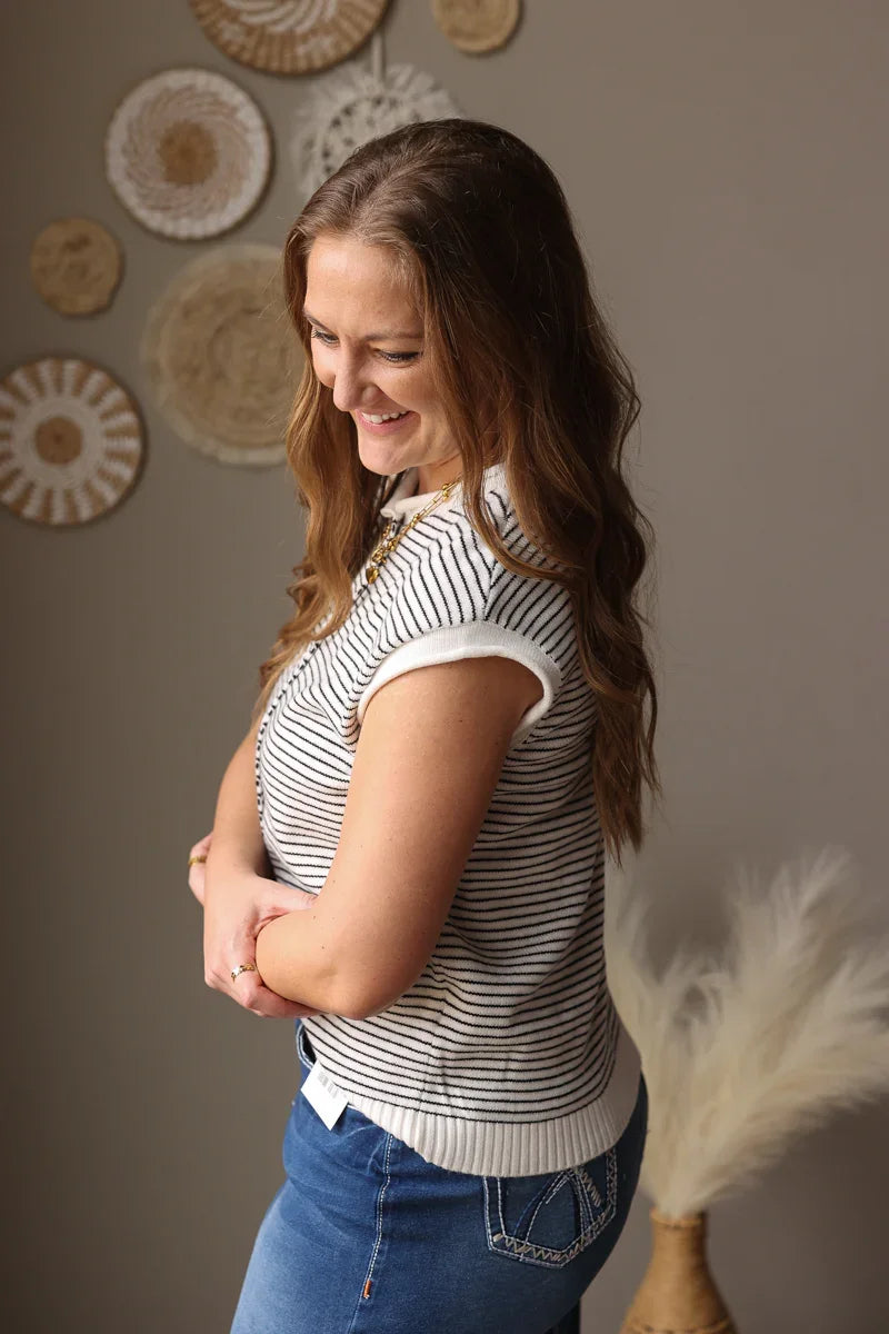 Woman wearing a striped shirt and blue jeans standing against a wall with decorative items.