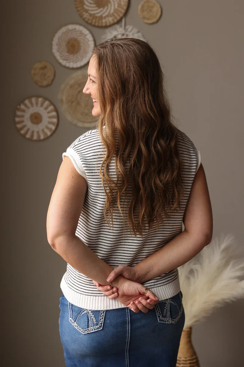 Woman standing with hands clasped, wearing a striped shirt and jeans, against a wall with decorative circular objects.