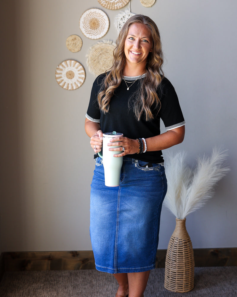 A woman stands indoors wearing a black short-sleeve top with white trim and a blue denim skirt, holding a white tumbler. The background shows a gray wall with decorative woven wall art and a woven vase with pampas grass.