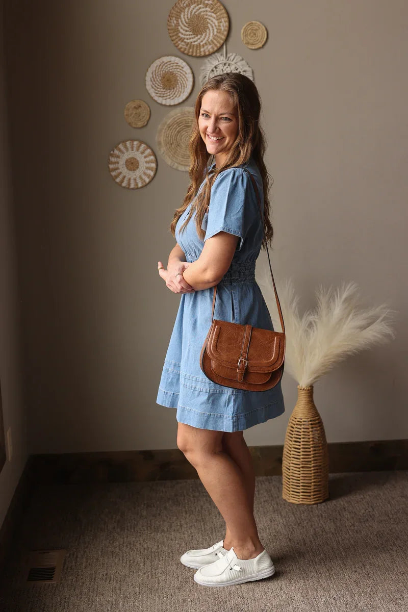 Woman in a blue dress with a brown bag standing in a room with decorative wall art and a vase.