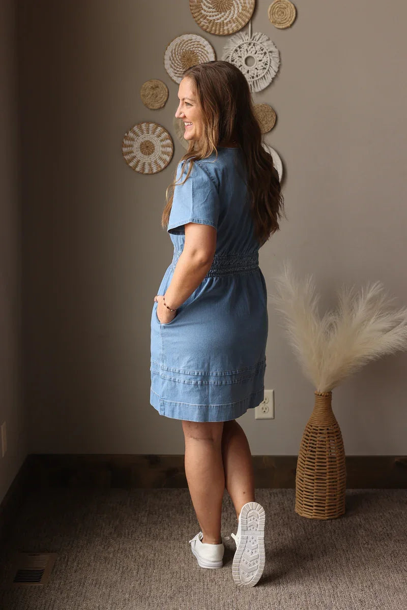 Woman in a blue dress standing in a room with decorative wall art and a vase.