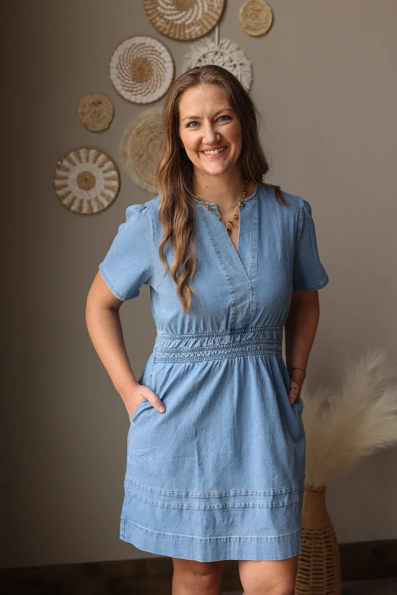 Woman wearing a blue dress standing against a wall with decorative items.