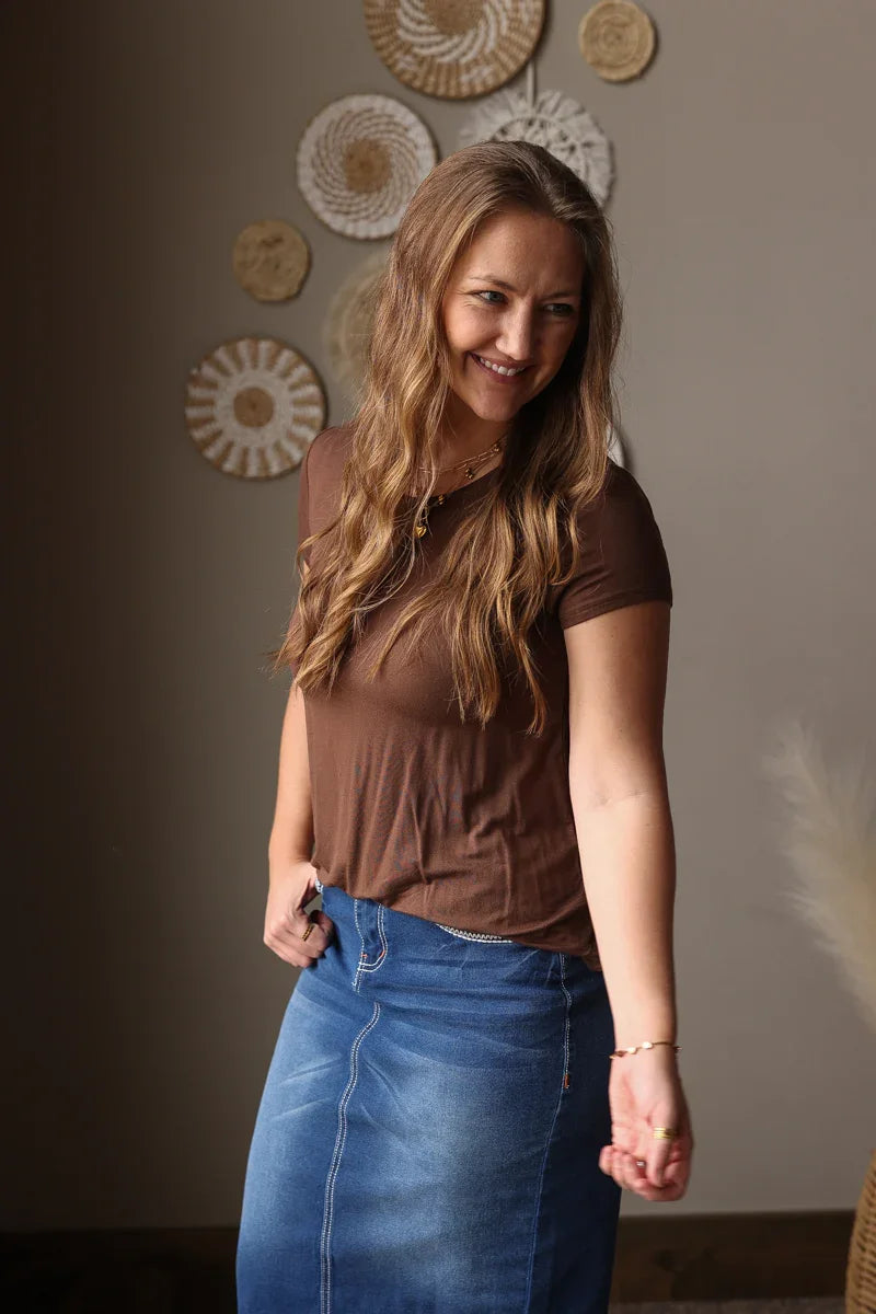 Woman wearing a brown top and blue jean skirt standing against a wall with decorative plates.
