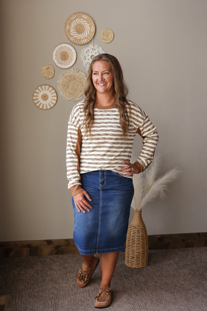 A woman modeling a beige and white striped long-sleeve top with a blue denim midi skirt, standing in a room with a neutral beige wall decorated with woven wall art and a pampas grass vase.