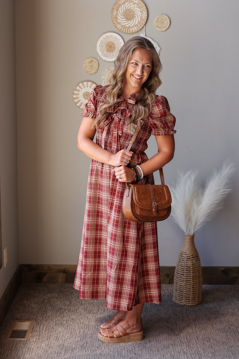 A woman wearing a red and brown plaid midi dress with puff sleeves and a ruffled tie neckline, holding a brown leather shoulder bag. She is standing in a room with a gray wall decorated with circular woven pieces and a vase of pampas grass.