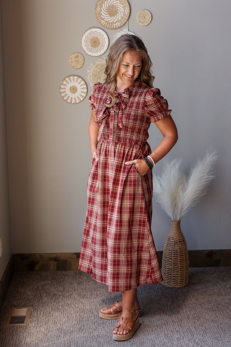 A woman modeling a red and white plaid midi dress with ruffled short sleeves and a tie-neck detail, standing in a styled room with a gray wall, decorative woven wall pieces, and a vase with pampas grass.